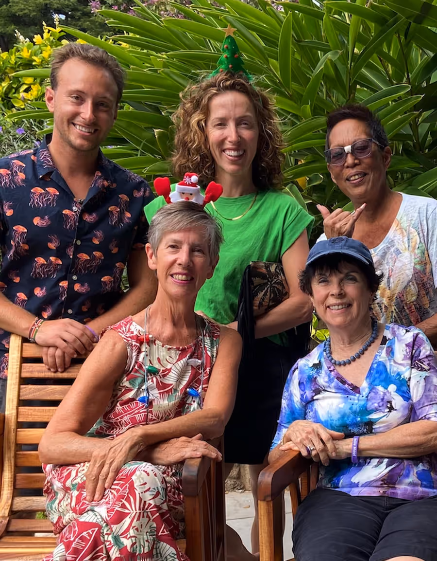 Group of five smiling adults, two seated in wooden chairs wearing festive accessories, with lush green plants in the background.