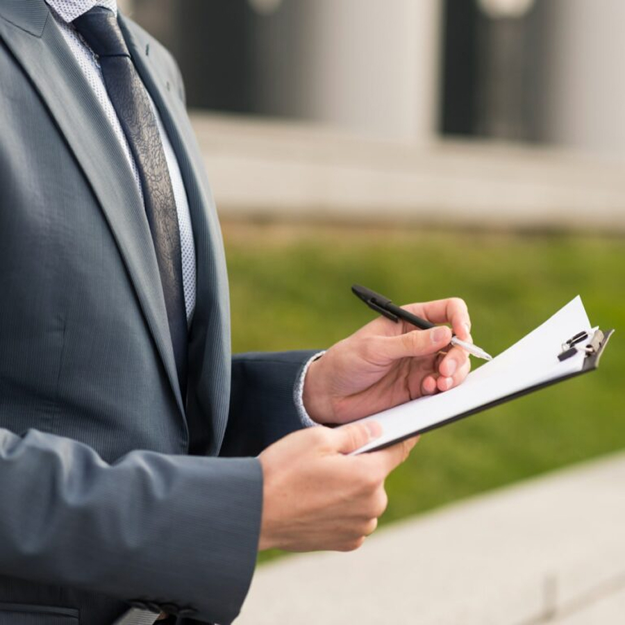 Person in a grey suit holding a clipboard and pen, outdoors near green grass.