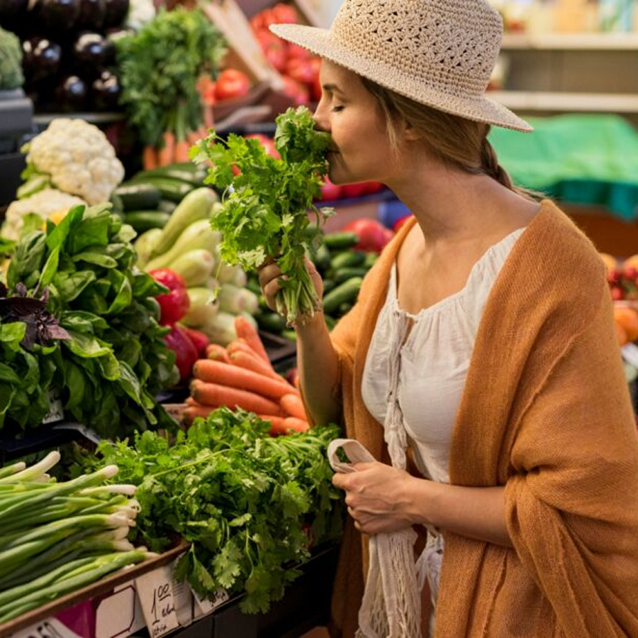 Woman wearing a hat smelling fresh cilantro in a grocery store produce section with various vegetables around.