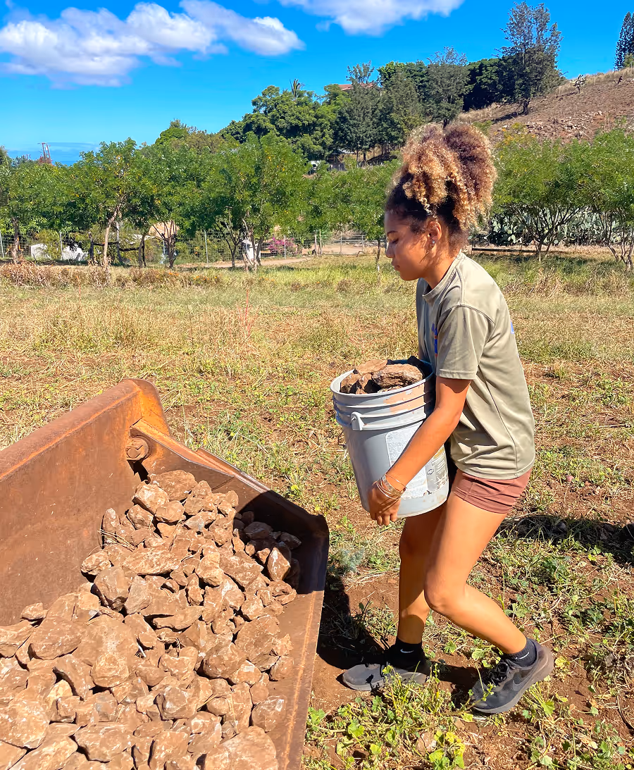 Young woman with curly hair carrying a bucket of rocks outdoors on a sunny day near a pile of rocks in a metal container.