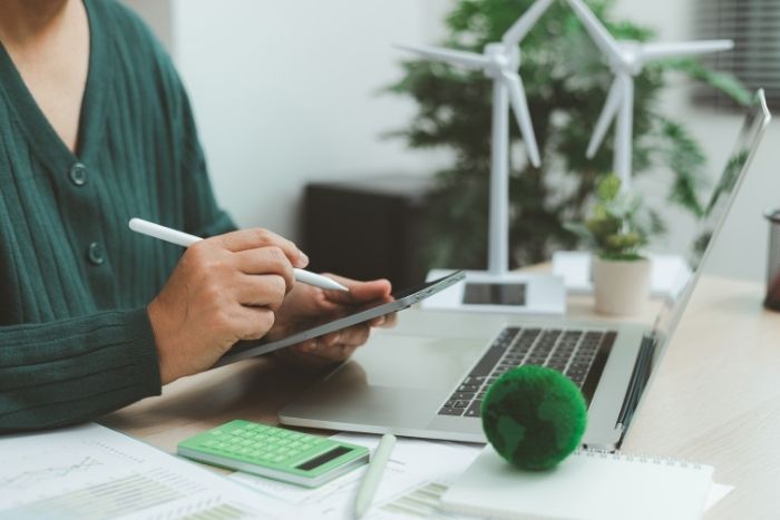 person sitzt am Schreibtisch und arbeitet. HĂ€lt ein Tablet in der Hand und vor der Person steht ein Laptop, Unterlagen und ein Taschenrechner.