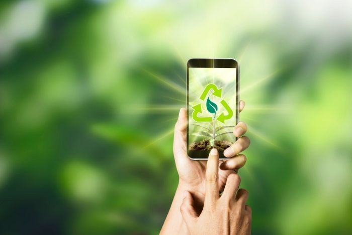 HĂ€nde halten ein Handy hoch auf dessen Display ein Recycling Zeichen grĂŒn strahlt.