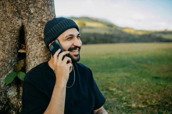 Ein Mann telefoniert, hält dafür ein Handy in der Hand und am Ohr und lehnt dabei an einem Baum.