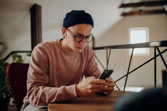 Junger Mann sitzt auf Stuhl an einem Tisch und hät ein Handy in seinen Händen.