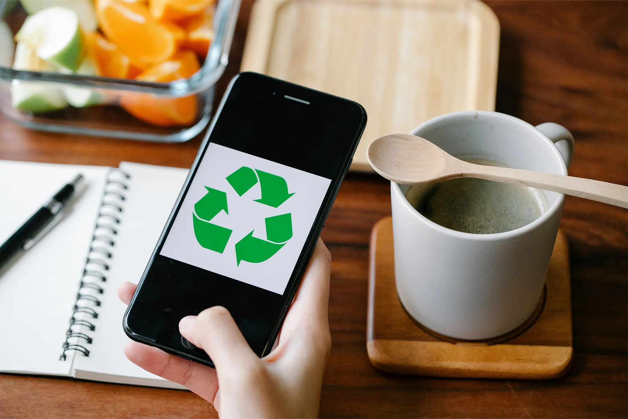 Smartphone mit Recycoling-Logo auf dem Bildschirm wird in der Hand gehalten und im Hintergrund sieht man einen Tisch mit Obst und einem Kaffee.