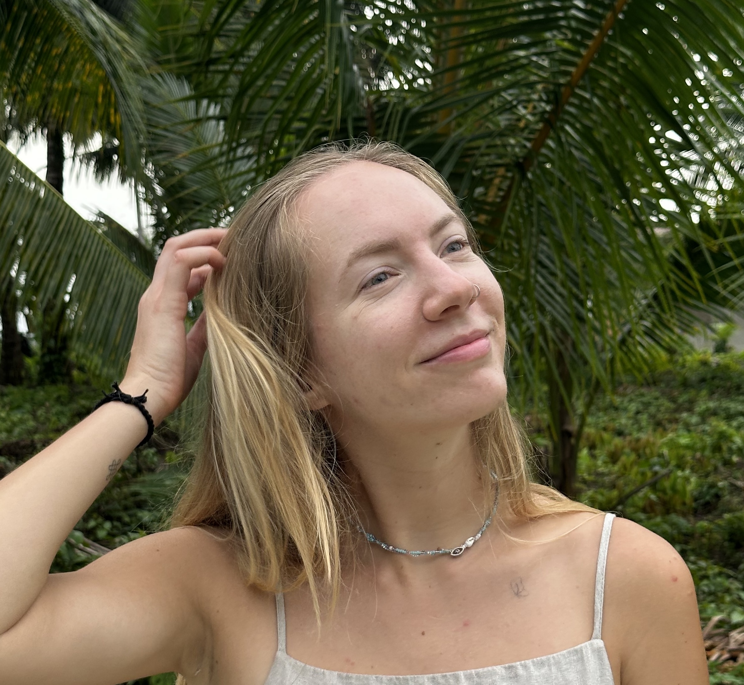 Young woman with blonde hair touching her hair, smiling slightly, standing outdoors with green palm leaves in the background.