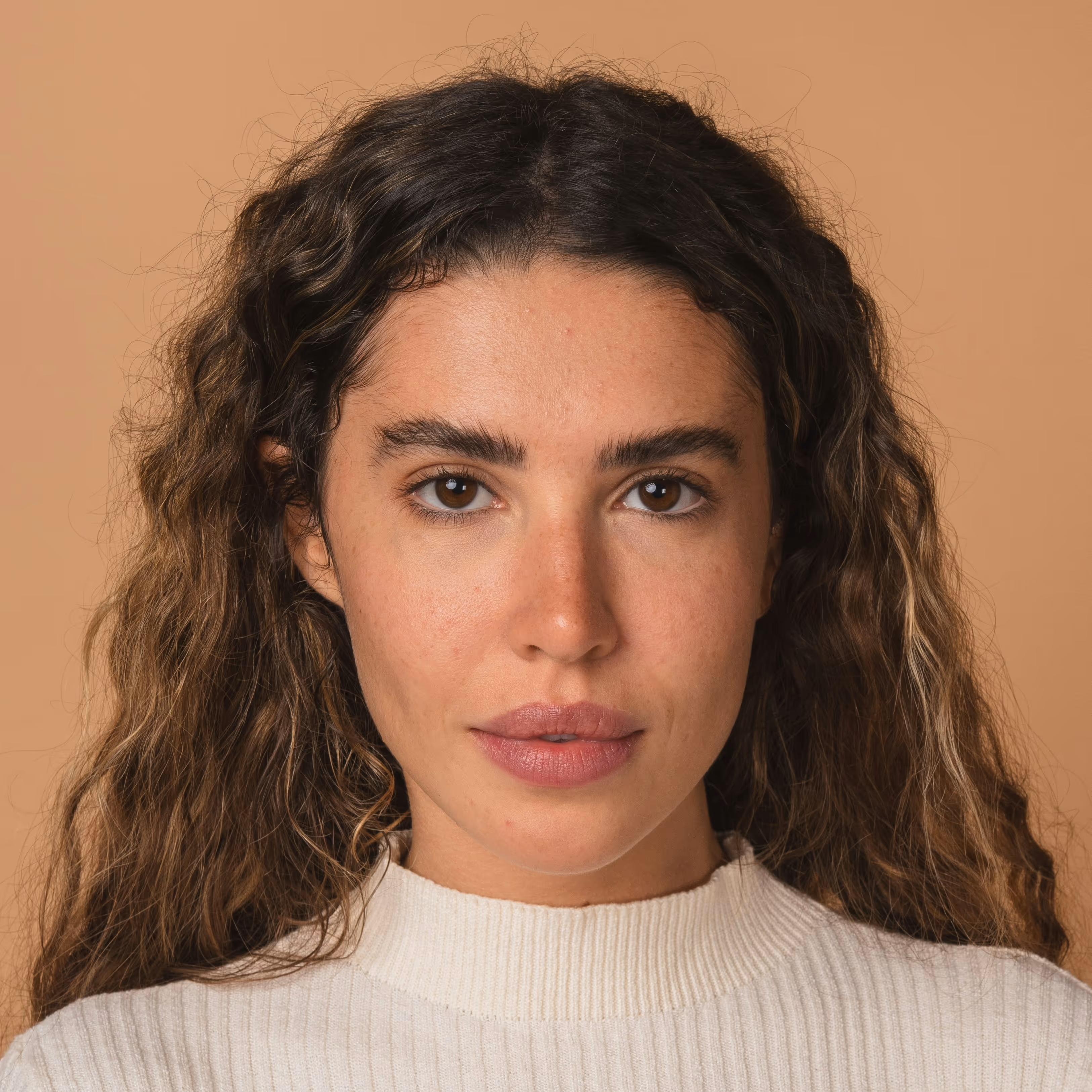 Close-up of a woman with long curly hair wearing a white ribbed sweater against a beige background.