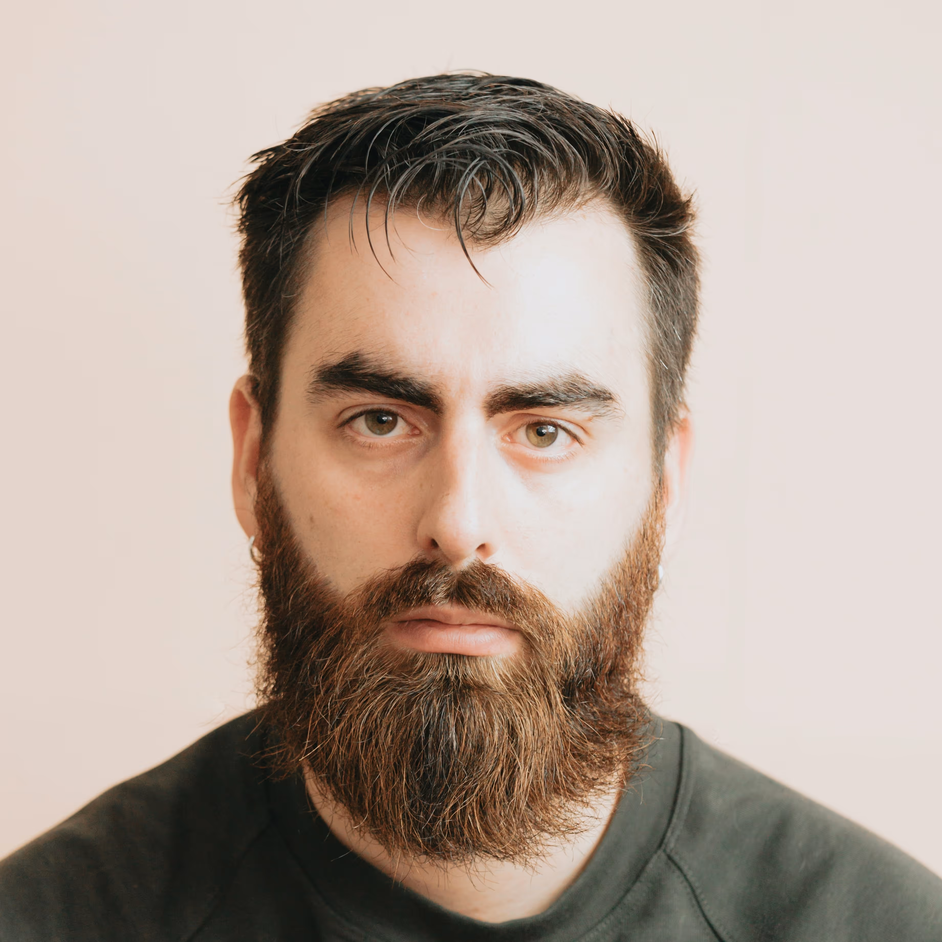Portrait of a serious man with dark hair, thick eyebrows, and a full beard wearing a dark shirt.
