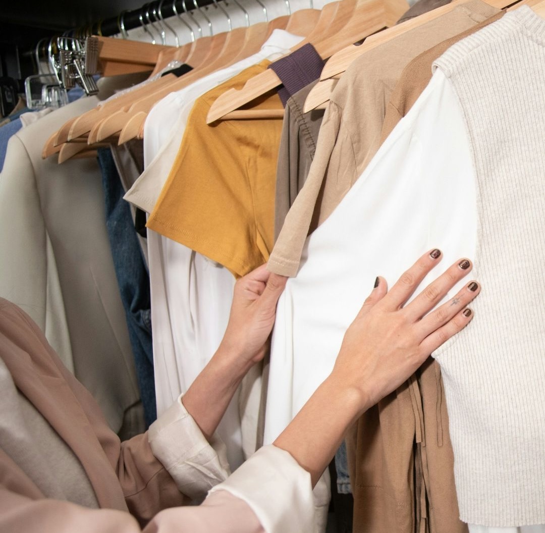 Hands browsing through a variety of neutral-colored shirts on wooden hangers in a wardrobe.