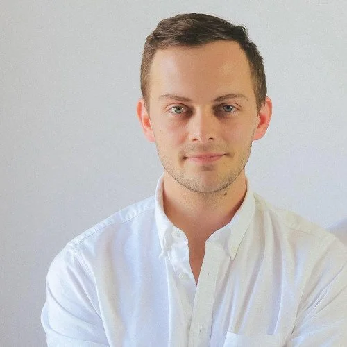 Young man with short brown hair wearing a white button-up shirt against a plain light background.