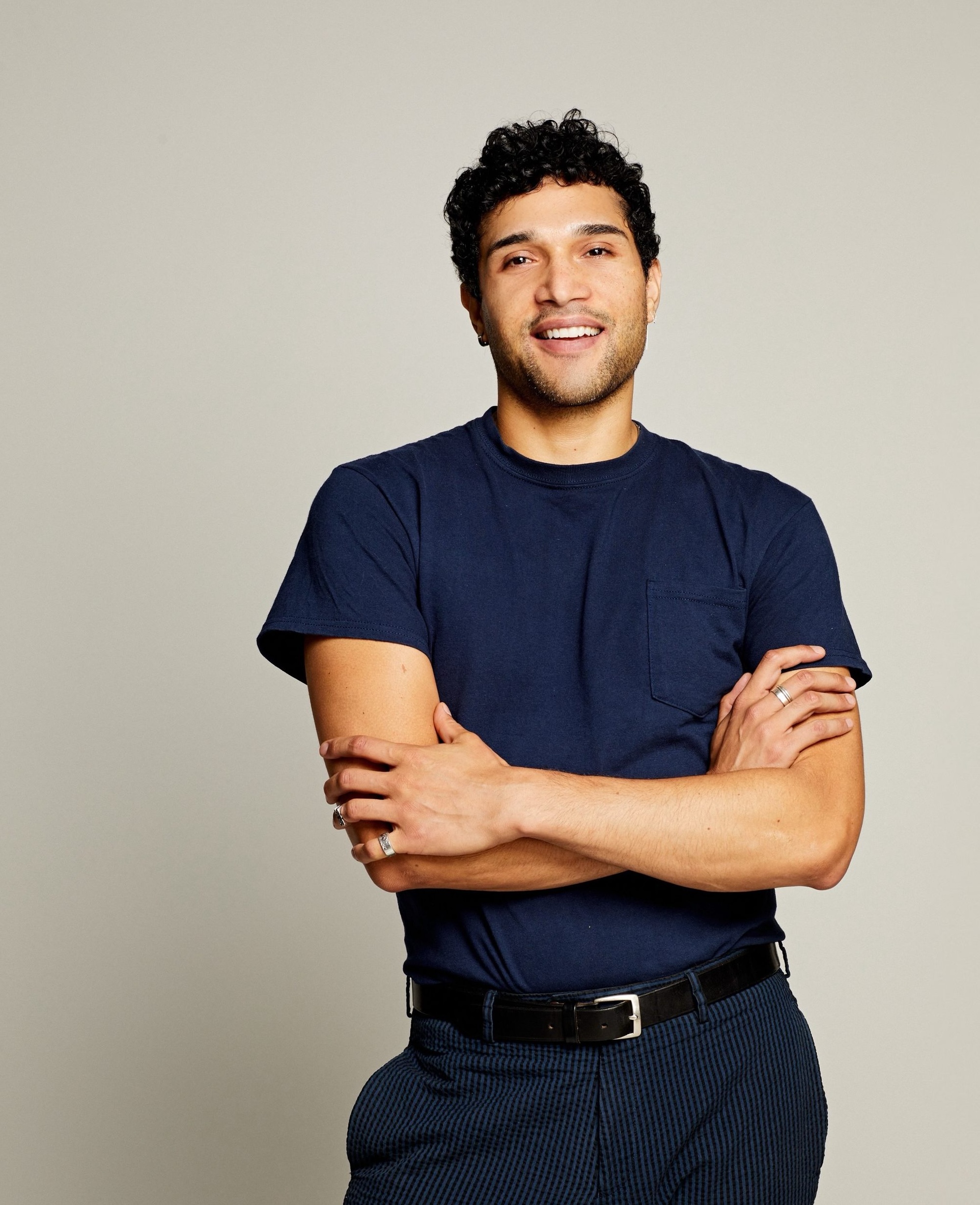 Smiling man with curly hair wearing a navy blue t-shirt and checkered pants standing with arms crossed.