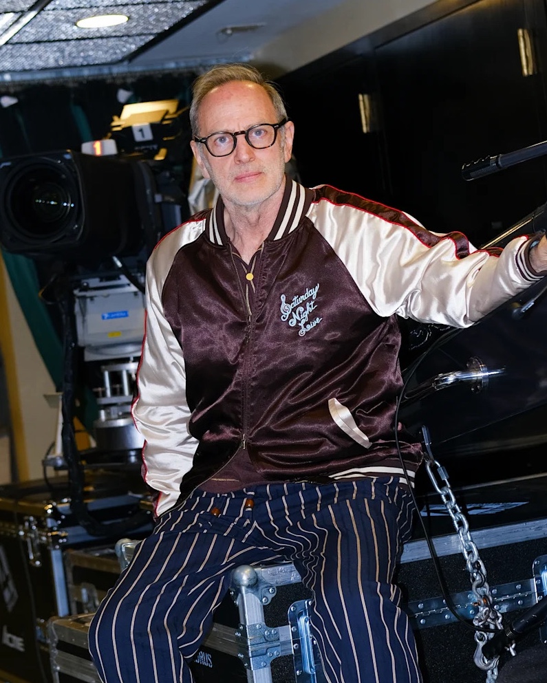 Man with glasses wearing a satin jacket with Saturday Night Live logo sitting on equipment cases.