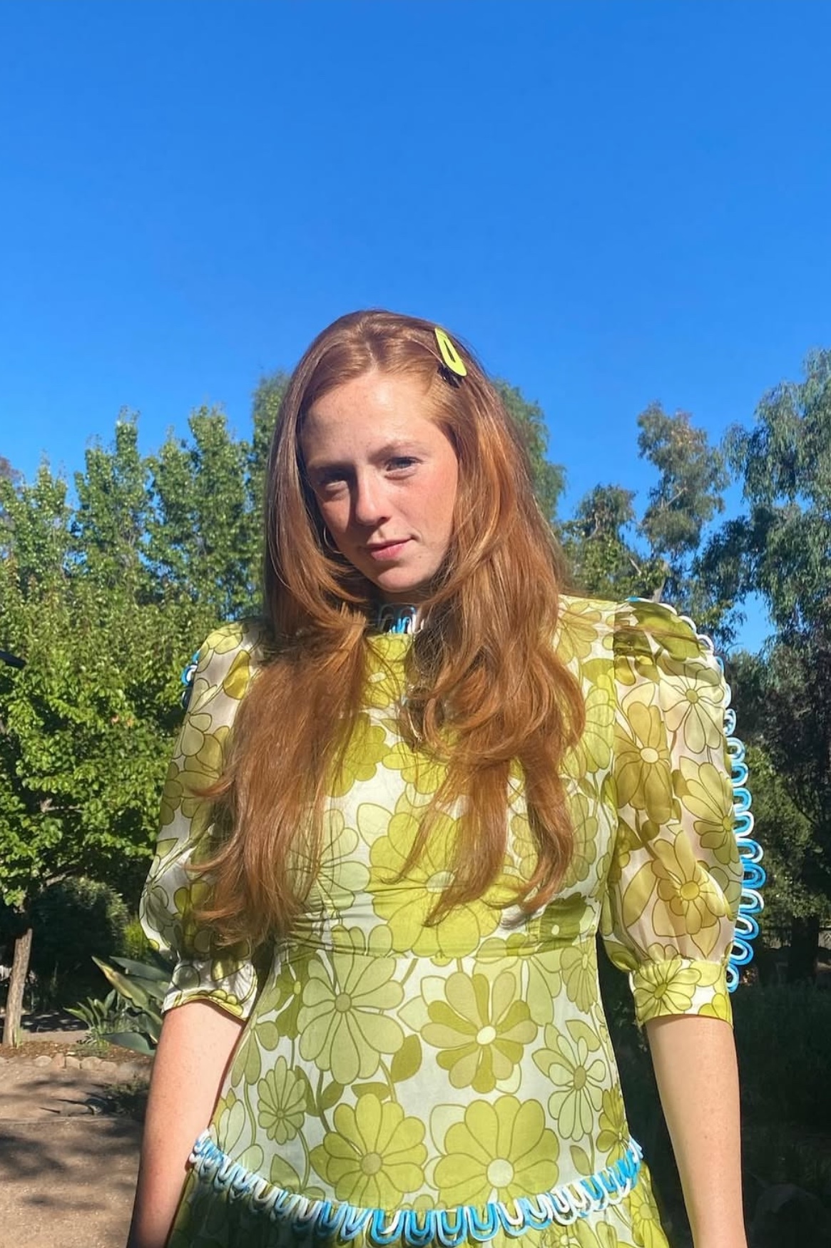 Woman with long red hair wearing a floral green and yellow dress standing outdoors under a clear blue sky.