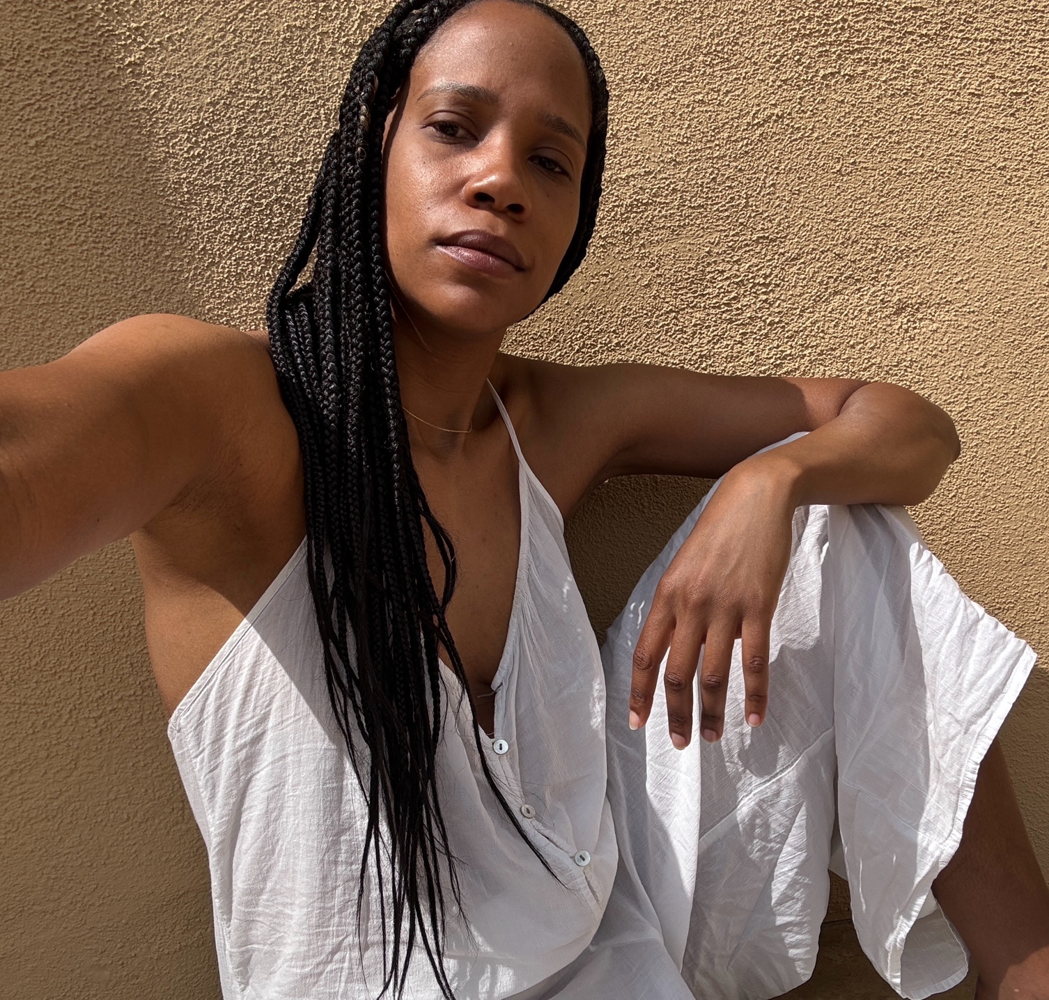 Woman with long braids wearing a white sleeveless dress sitting against a textured beige wall.