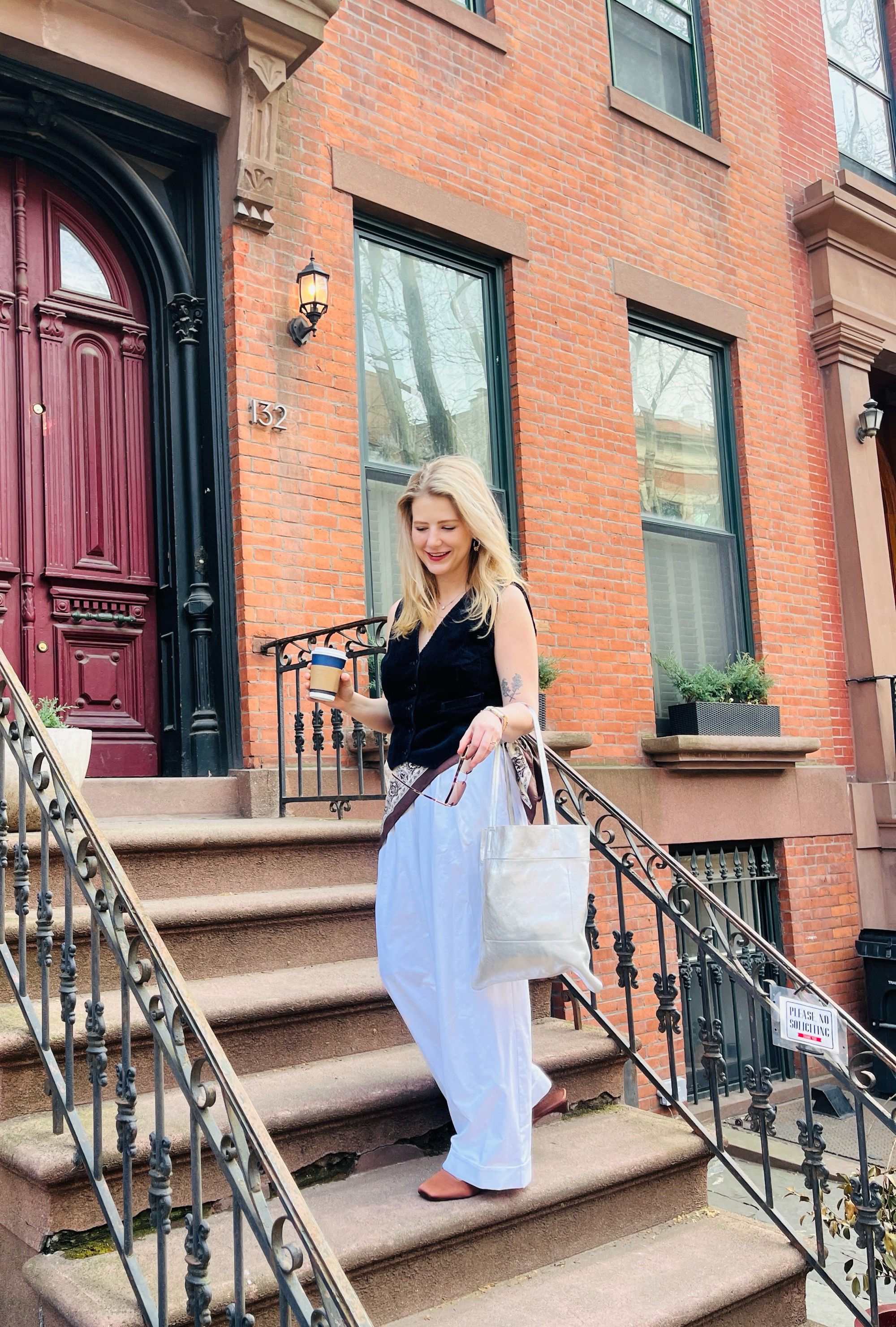 Woman with blonde hair wearing a black vest and white pants stands on brownstone steps holding a coffee cup and white bag.