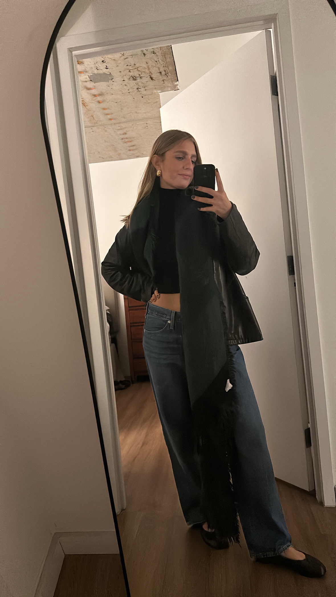 Woman taking a mirror selfie wearing a black cropped top, leather jacket, scarf, wide jeans, and flats.