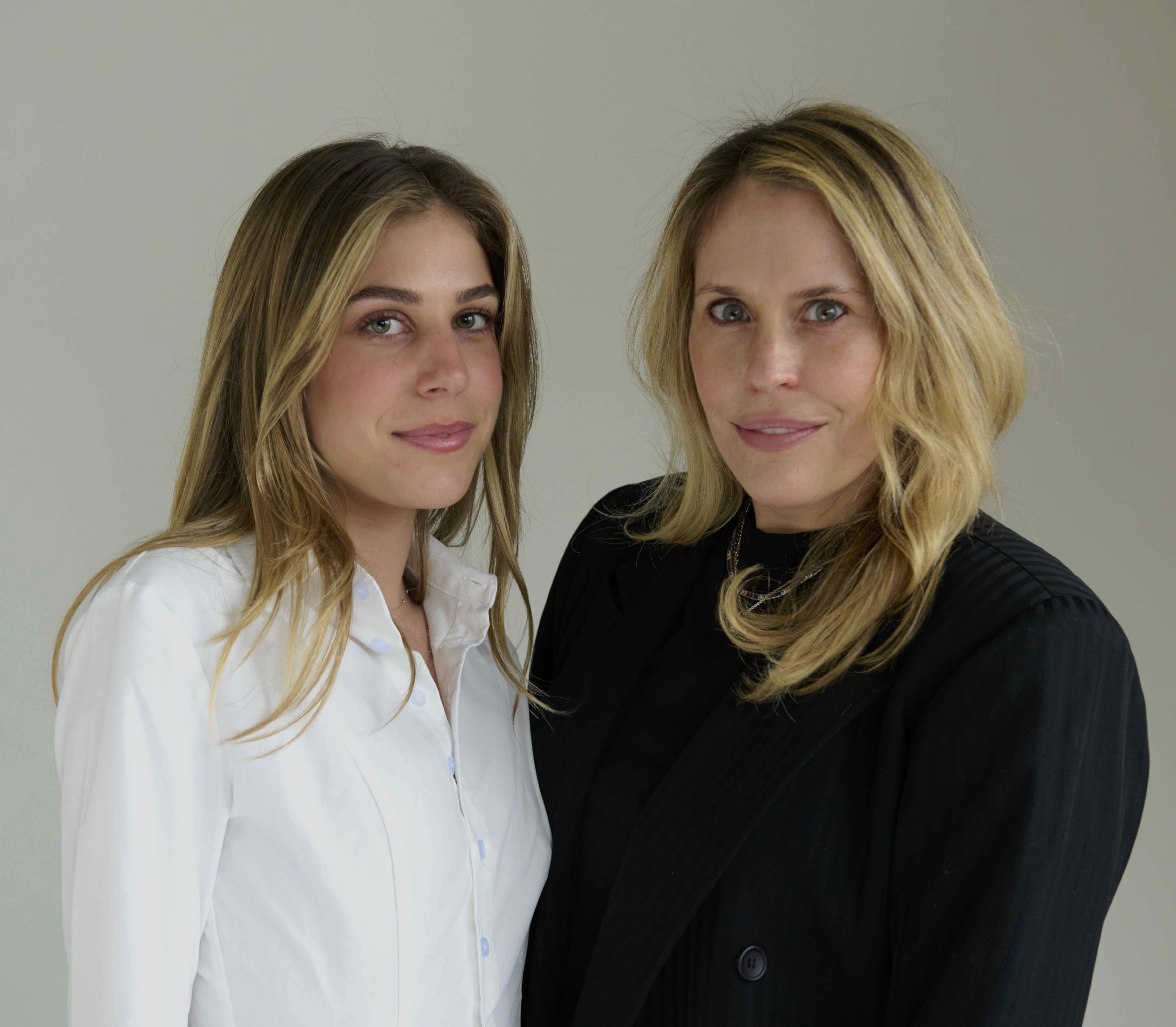 Two blonde women posing against a plain gray background, one in a white shirt and the other in a black outfit.