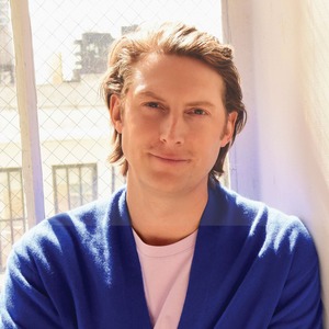 Person with light brown hair wearing a blue cardigan and light pink shirt in a sunlit room.