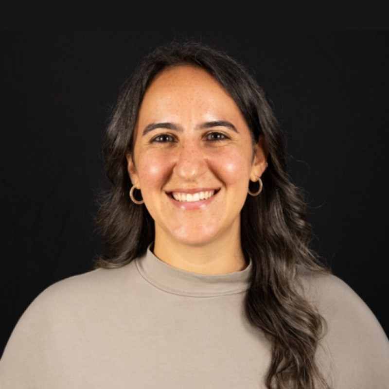 Smiling woman with long dark hair and hoop earrings wearing a light gray top against a black background.