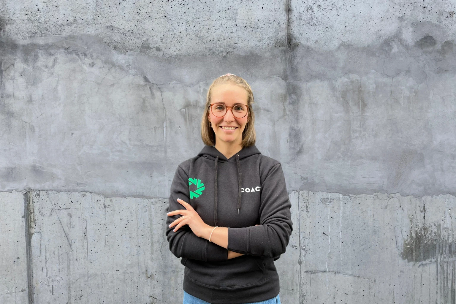 Junge Frau mit Brille und grauer Kapuzenjacke steht vor Betonwand und lächelt in die Kamera.