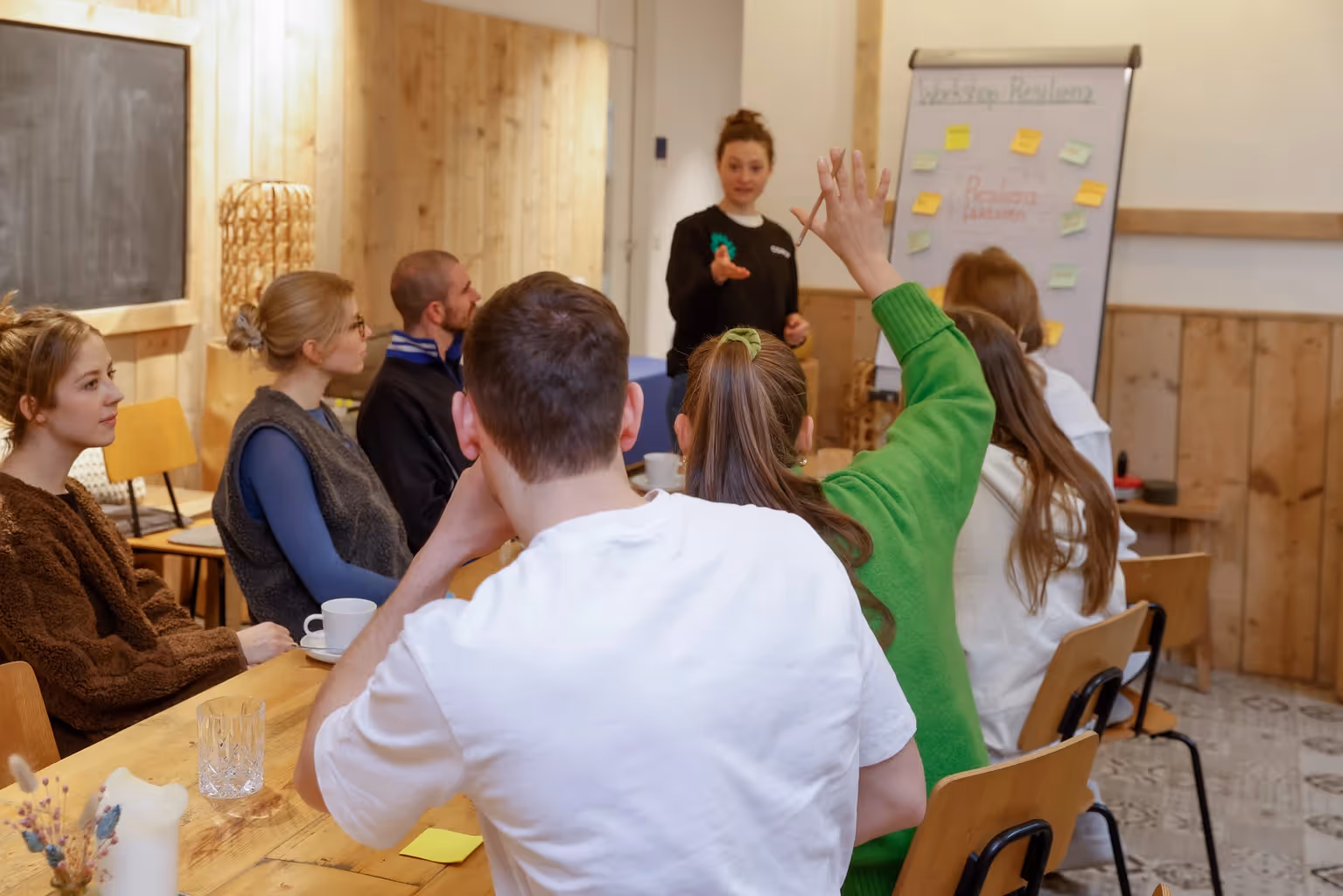 Ein Team sitzt gemeinsam an einem Tisch, eine Frau steht an einem Flipchart und eine Frau am Tisch meldet sich