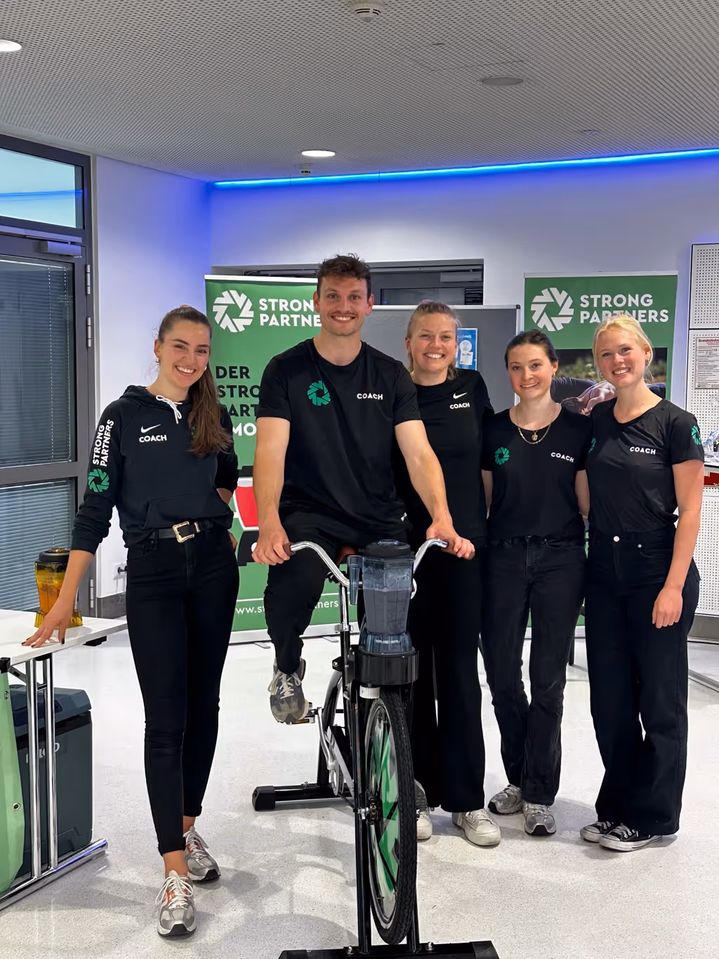 Srong Partners Coaches stehen zusammen und ein Coach sitzt auf dem Smoothie Bike