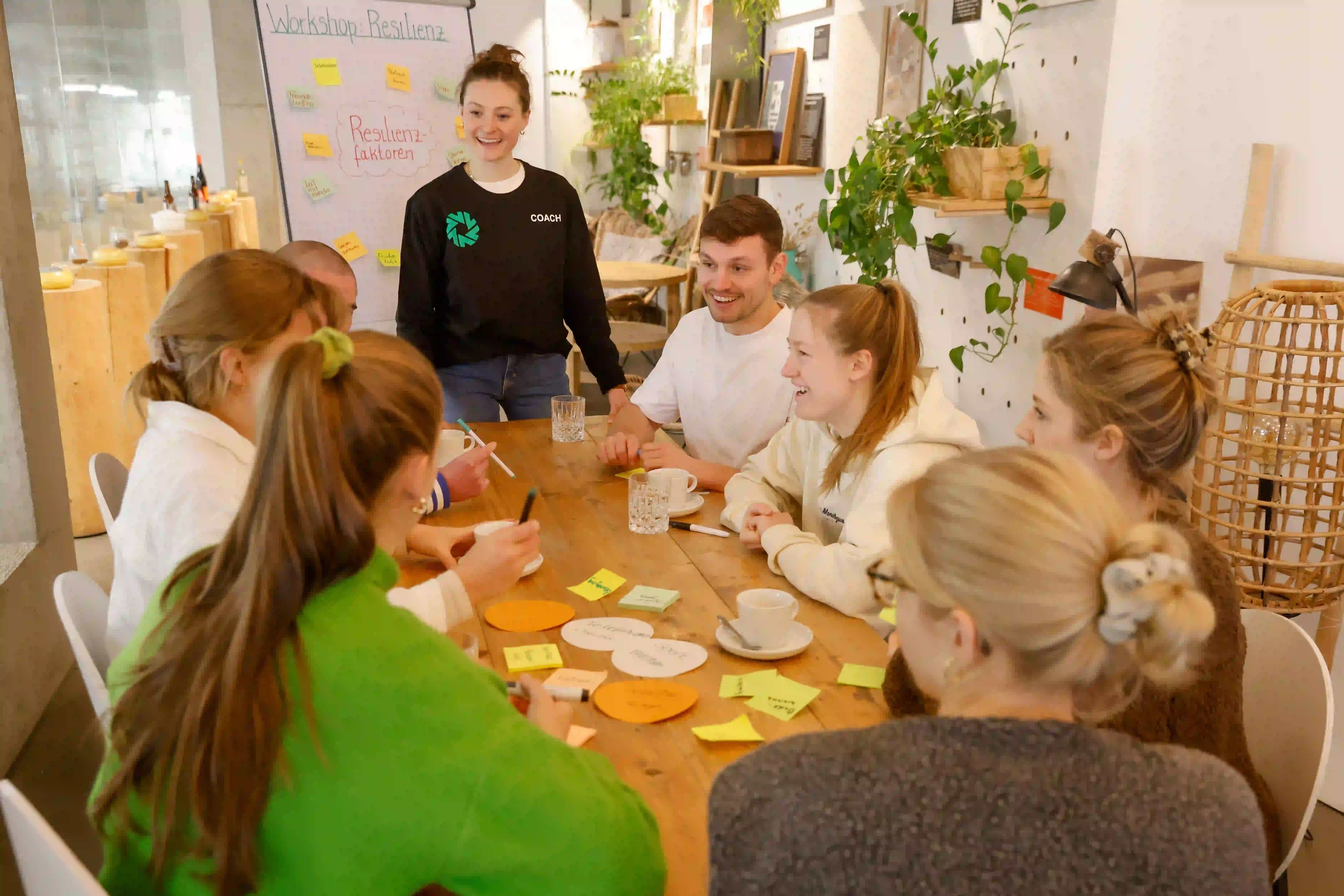 Workshop Situation mit Teilnehmenden an einem Tisch und Strong Partners Coach steht am Flipchart