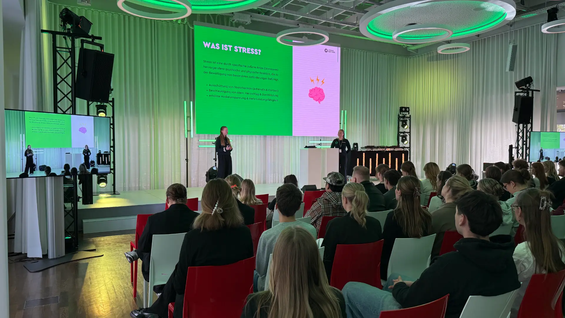 Zwei Referentinnen von Strong Partners halten vor vollem Saal einen Workshop zur Stressregulation, auf der Leinwand die Folie „Was ist Stress?“