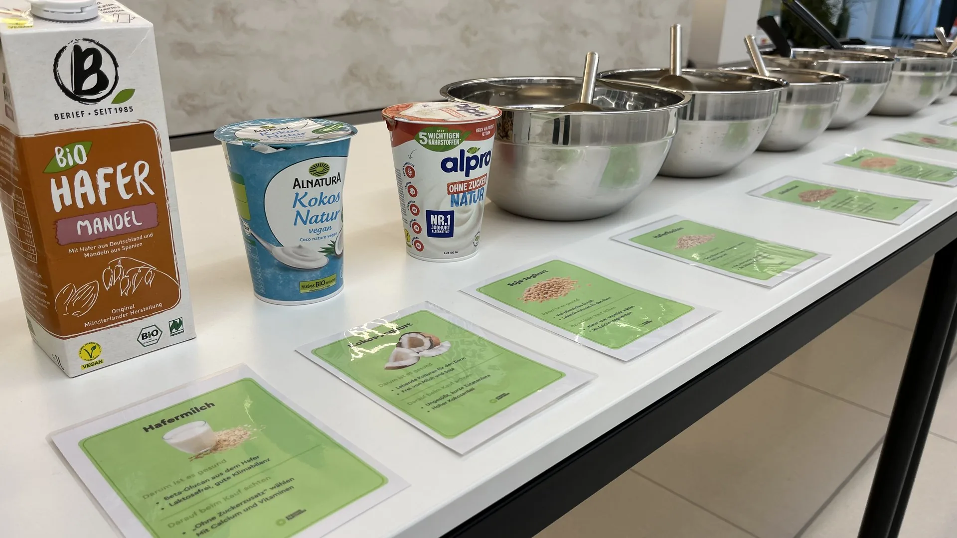 Frühstücksbuffet beim How to Müsli Workshop mit Haferdrink, pflanzlichen Joghurts, Edelstahlschüsseln und grünen Infokarten zu den Zutaten.