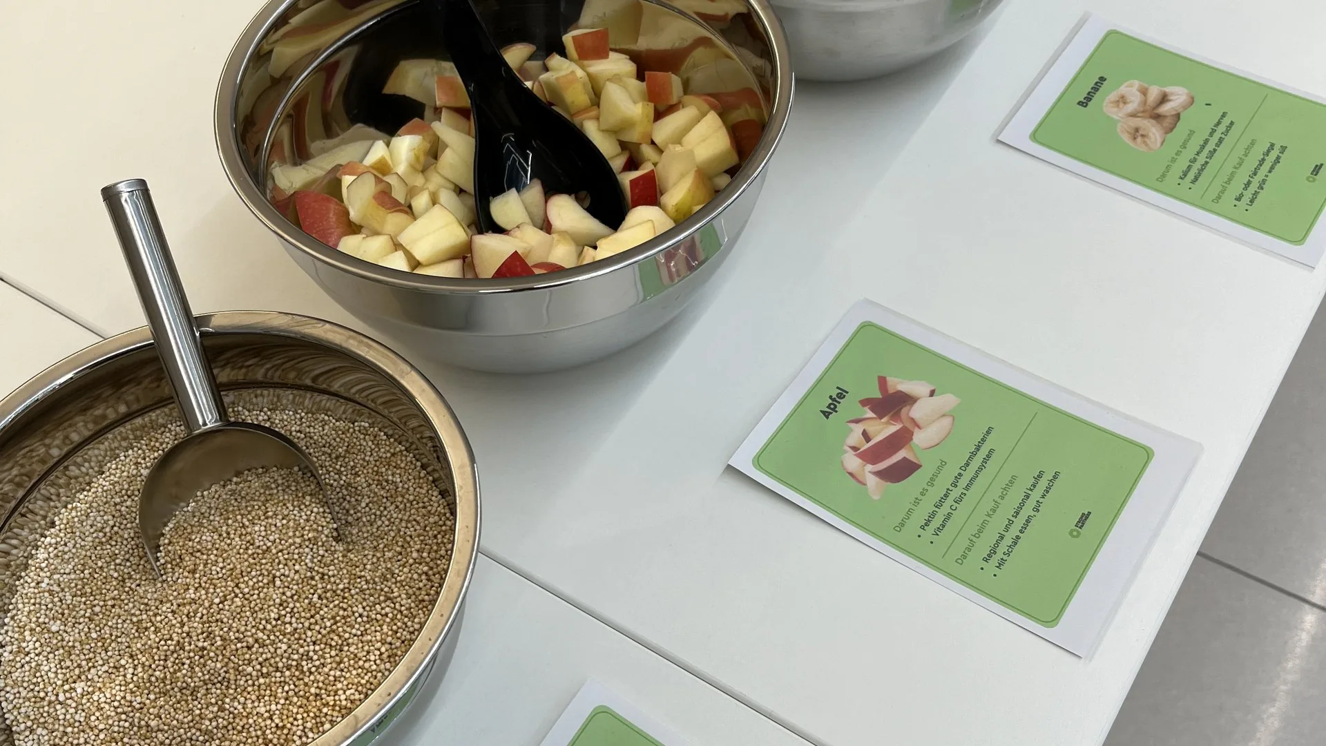 Schüsseln mit Amaranth, Apfelstücken und Bananenscheiben am Müsli-Buffet, davor Infokarten zu jeder Zutat