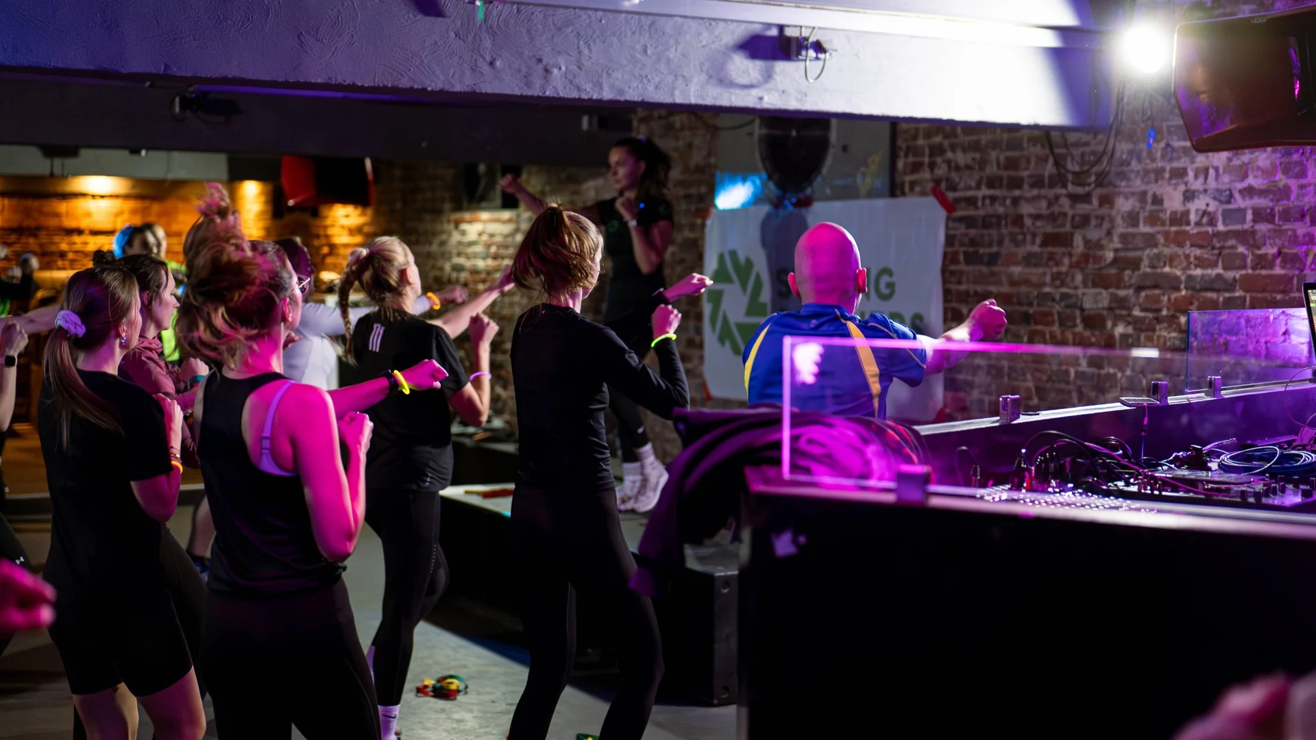 Gruppe beim Fitnesskurs mit Trainerin in einem Club mit Backsteinwänden und lila Licht, im Hintergrund ein Strong-Partners-Banner.