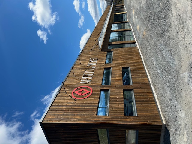 Moderne bygning med trepanel og vinduer under blå himmel med skyer, med logo og skilt som sier Arbeid og Vekst.