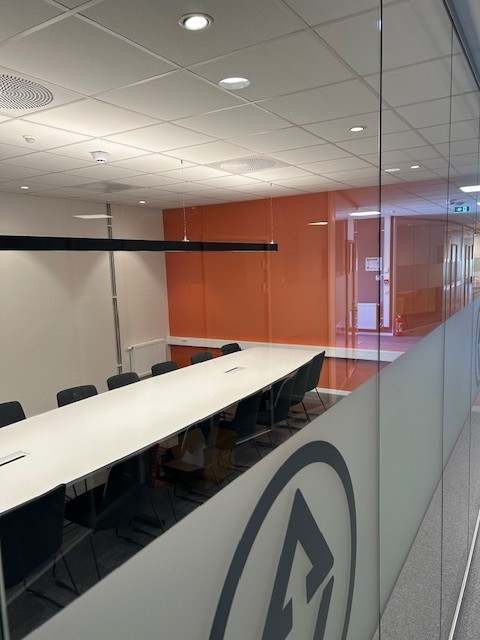 Moderne møterom med et langt hvitt bord, flere svarte stoler, oransje vegg og glassvegg med logo.