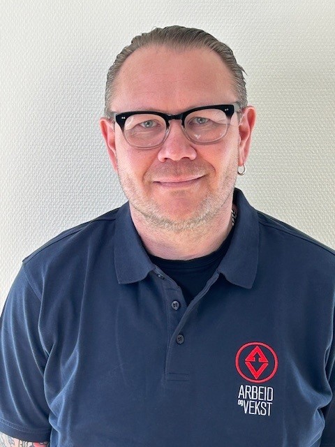 Mann med briller og blå skjorte med Arbeid og Vekst-logo smilende mot kamera.