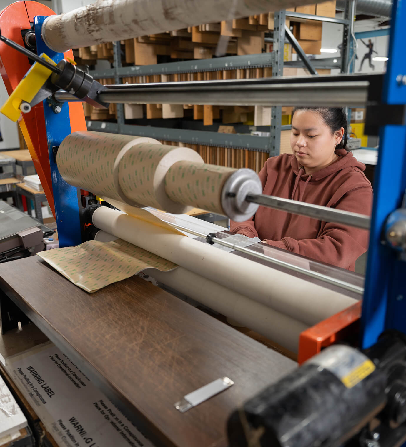 A production worker feeding material through a wide roll laminator loaded with 3M adhesive