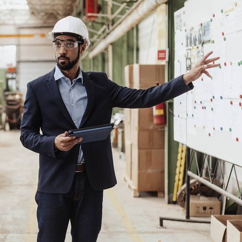 A manager in a hard hat reviewing a production planning board in a warehouse