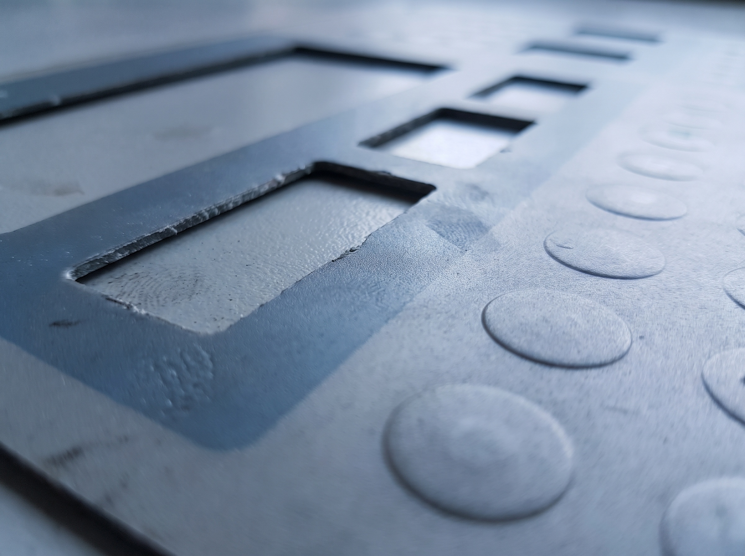 Raking light across a control panel overlay, showing die-cut window openings and rows of raised embossed key areas.