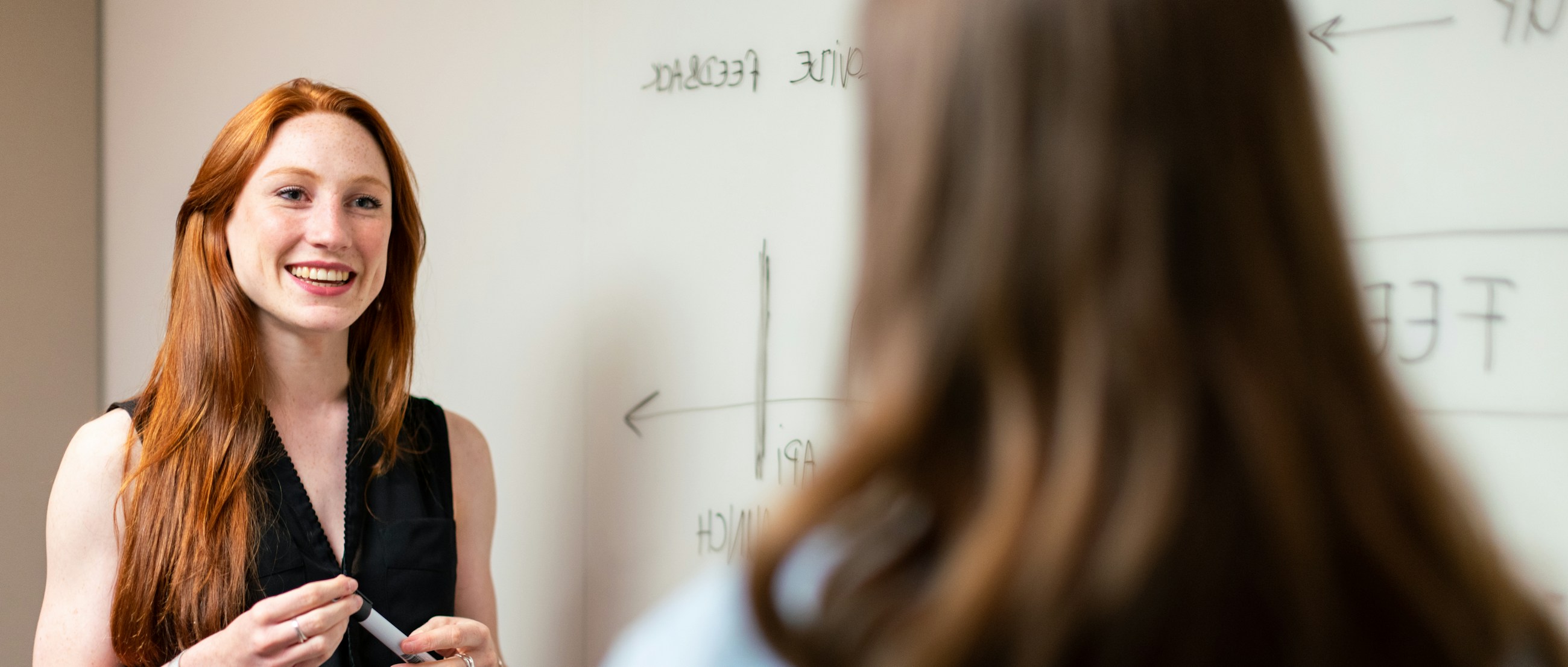 A smiling woman with long red hair in a black sleeveless top standing at a whiteboard