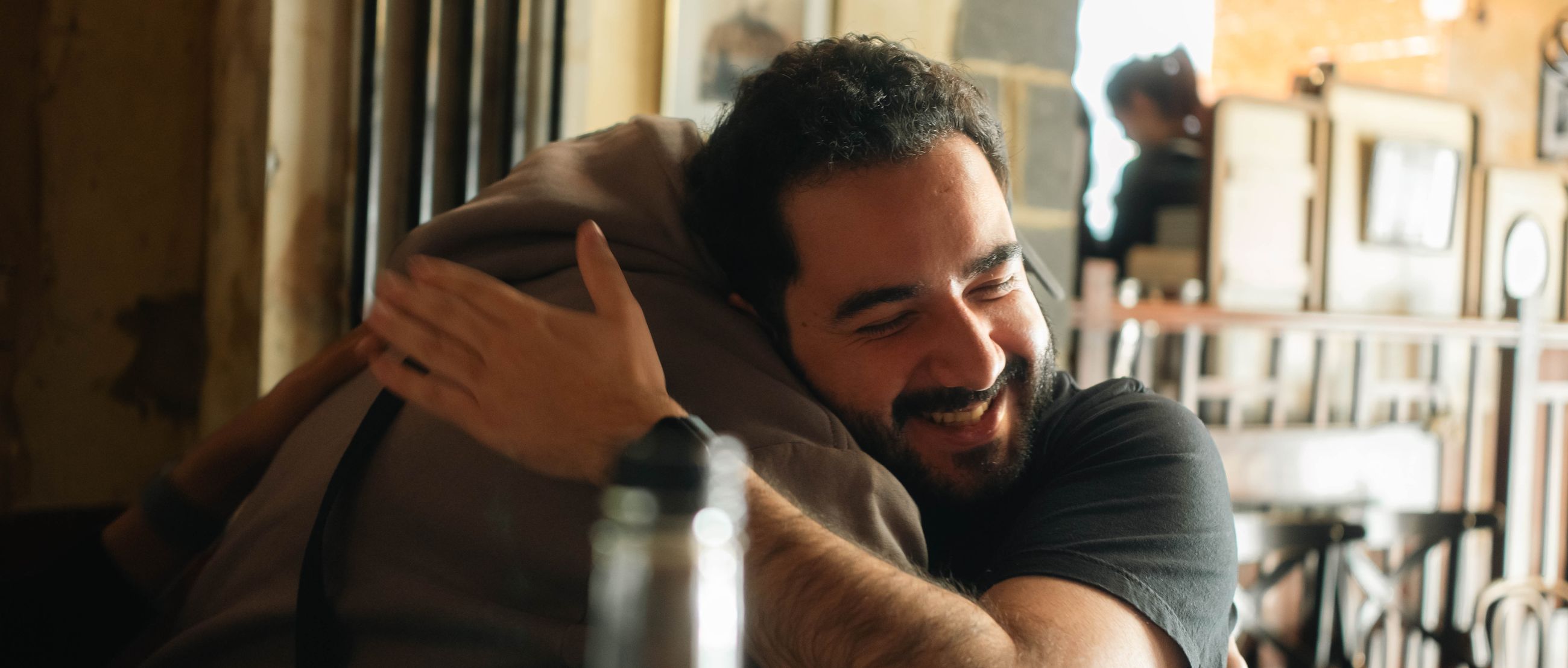 A man smiling softly with his eyes closed while embracing someone in a warm, sunlit room