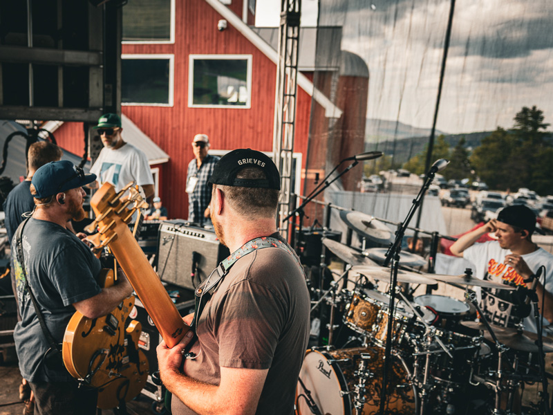 Band getting ready on an outdoor stage in front of a red barn, with mountains in the distance
