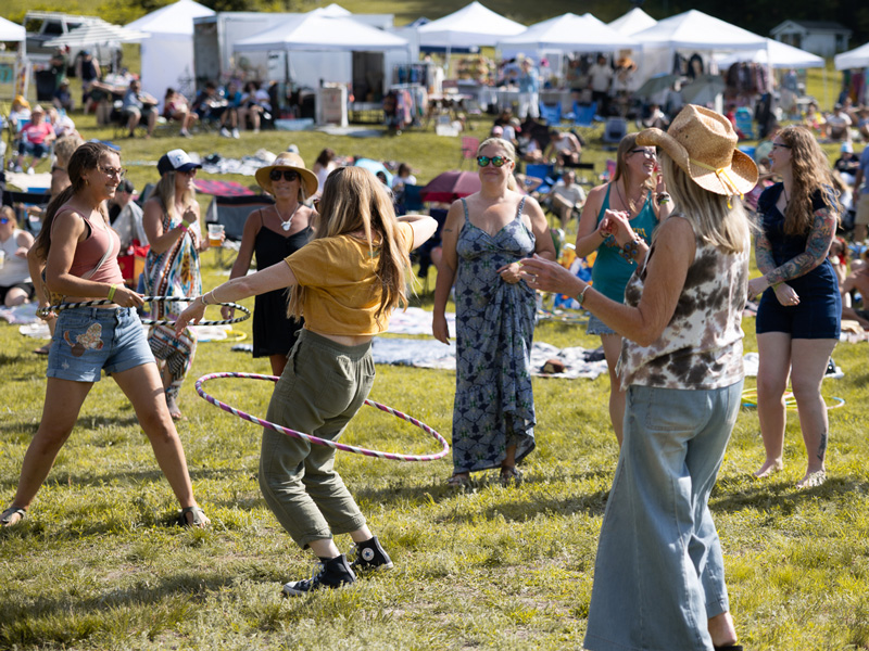 Festivalgoers hula hooping and dancing on the grass near white vendor tents