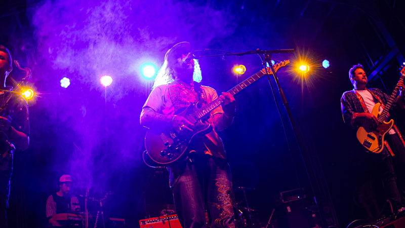 Long-haired guitarist singing through purple stage lights and haze with his band