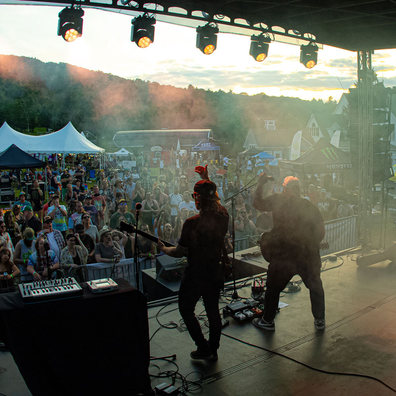 View from behind two guitarists on stage, looking out over a large crowd at sunset