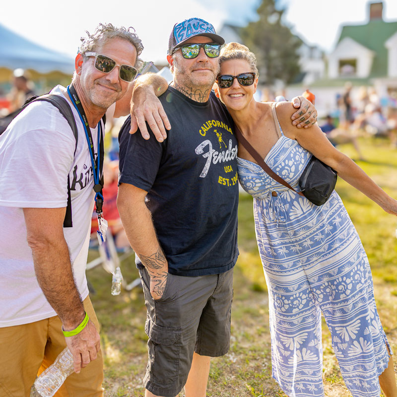 Three smiling festivalgoers in sunglasses posing arm in arm on a sunny day