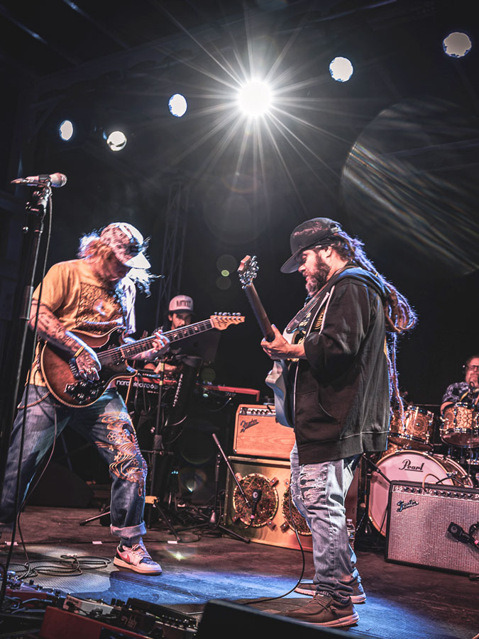 Two guitarists facing each other under bright stage lights, with a drummer behind them