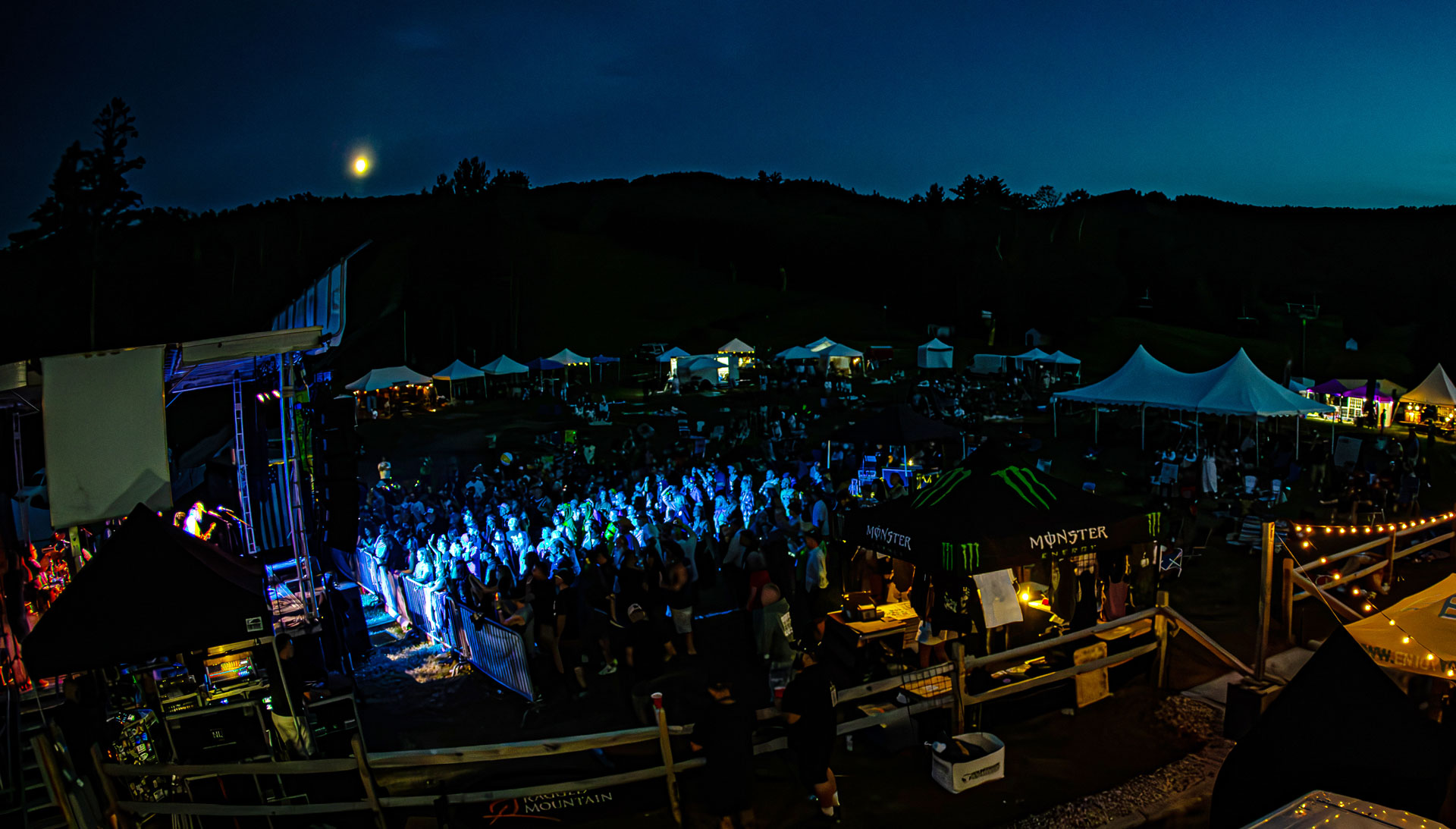 Crowd under blue stage lights at a nighttime Kinfolk concert, with vendor tents and a full moon over the hills
