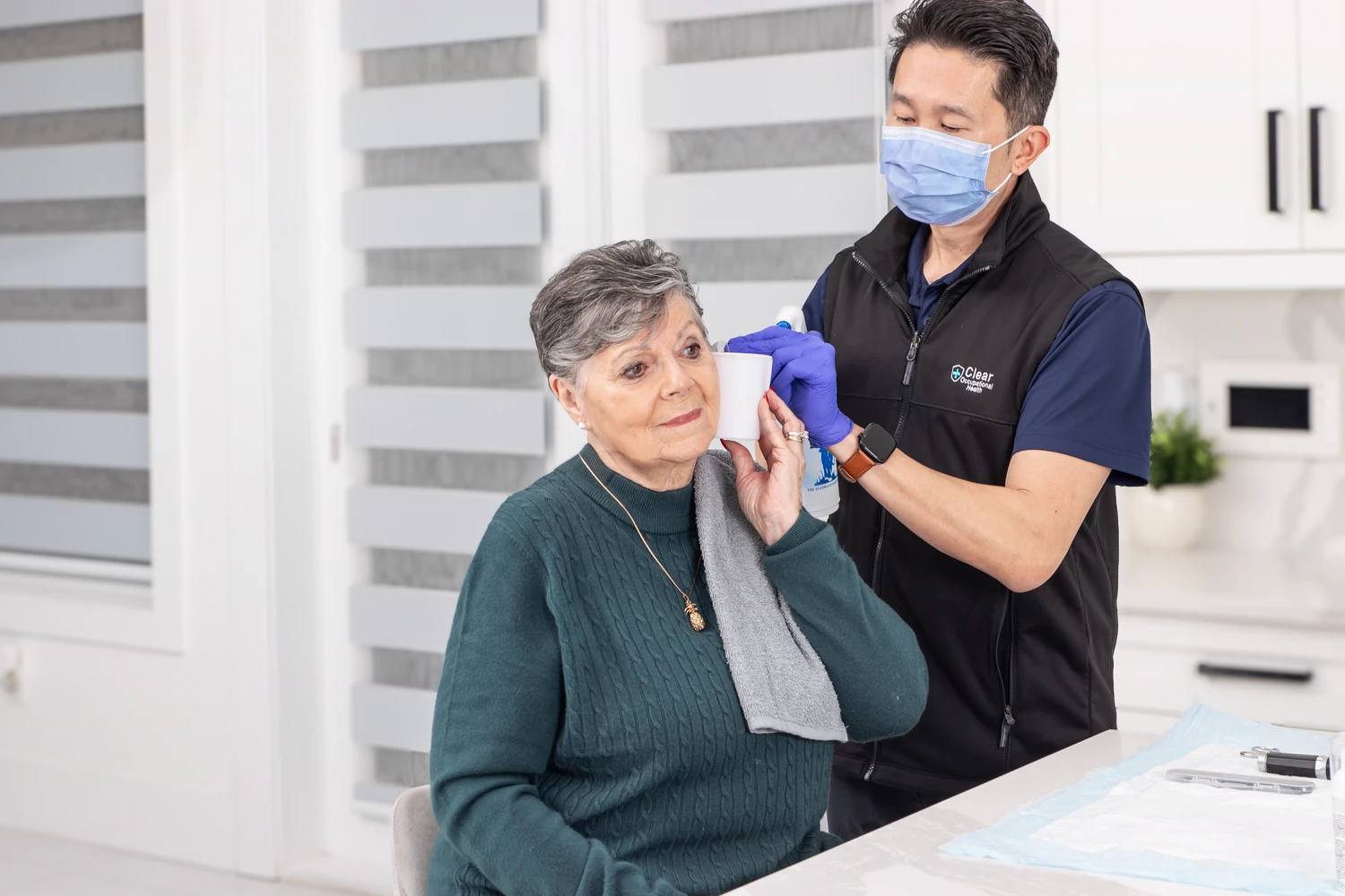 Masked technician performing ear irrigation for a senior woman at home in Coquitlam