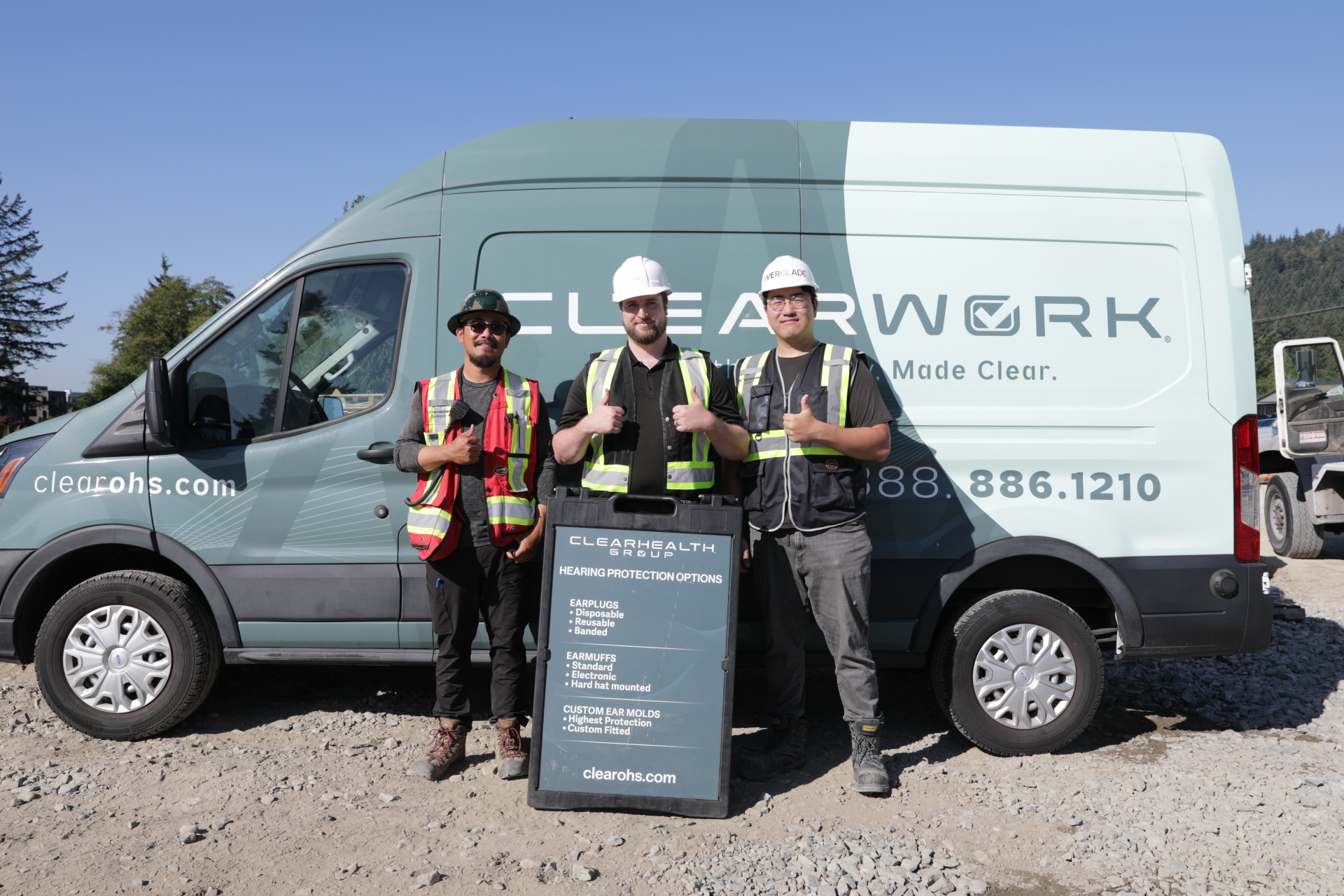 Three Clear Work technicians in hi-vis and hard hats beside one of the Clear Work mobile units at a B.C. worksite, holding custom-moulded hearing protection