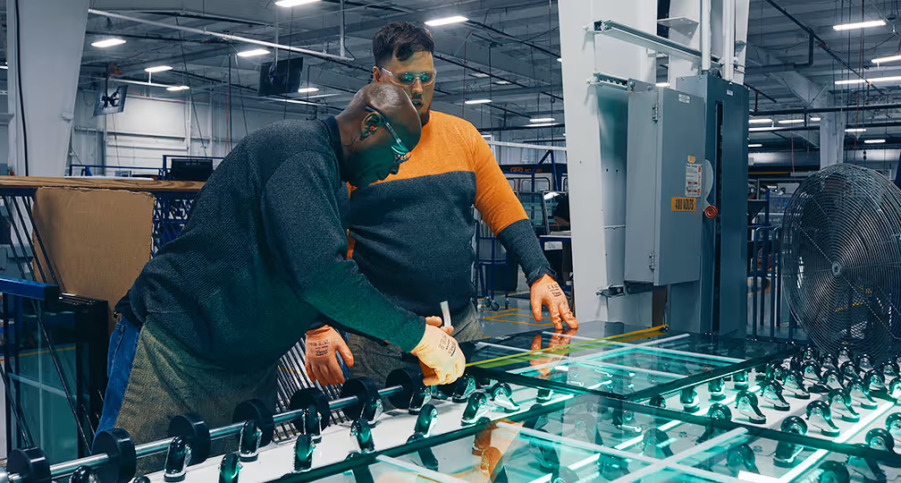 Two factory workers wearing safety glasses and gloves inspecting and measuring sheets of glass on machinery in an industrial setting.