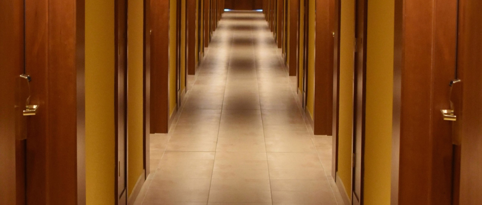 Long hotel hallway with multiple closed wooden doors on both sides and beige tiled floor.